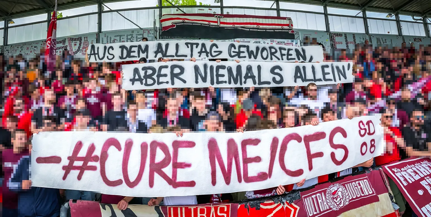 Photo d'une tribune de match de foot. Les supporter portent une bannière : "Cure ME/CFS" (Guérissez l'EM/SFC). Deuxième bannière en allemand : "Arraché à la vie quotidienne, mais jamais seul".
